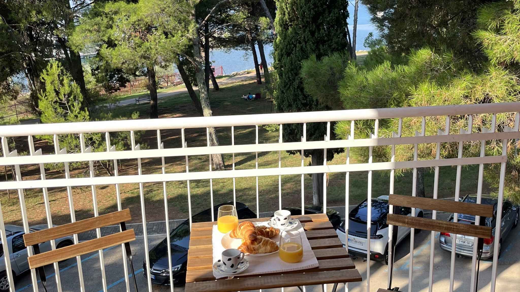 A balcony with a table set for breakfast, overlooking a park and a body of water.