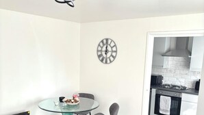 A modern living room with a glass table, grey chairs, a black sofa, and a kitchen visible through an open door. - Freshly Renovated 2 bed Near City Airport (London)