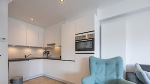 A modern kitchen with white cabinets, a built-in oven, and a coffee machine. A light blue armchair is placed in the living area. - Stylish apartment with terrace close to the sea (Middelkerke)