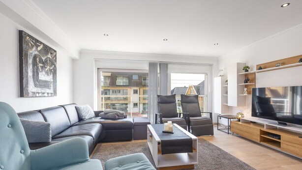 A modern living room with a sofa, armchairs, a coffee table, and a TV. There is a large window with a view of buildings outside. - Stylish apartment with terrace close to the sea (Middelkerke)