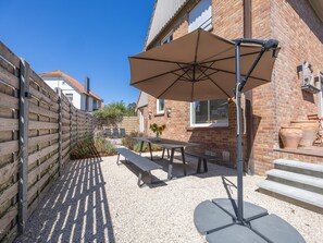 An outdoor patio area with a table and benches, a wooden fence, and a brick building in the background. - Luxury holiday home for 8 people near the sea (Westende)