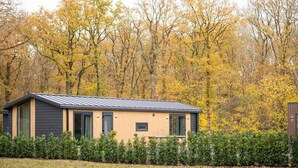 Eine moderne Hütte mit geneigtem Dach, großen Fenstern und einer Holzfassade, umgeben von einem gepflegten Garten und Herbstbäumen im Hintergrund.