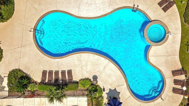 Vista aèria d'una piscina amb un disseny ondulat singular, rodejada de gandules i una zona d'aparcament.