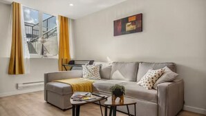 Ein modernes Wohnzimmer mit einem grauen Sofa, einem Couchtisch und einem kleinen Beistelltisch. Ein Fenster mit Vorhängen und ein Bild an der Wand sind zu sehen.