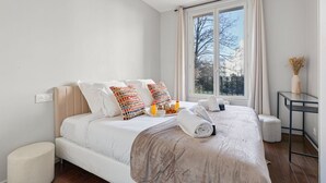 Quarto de hotel moderno com uma cama grande, uma mesinha de cabeceira, uma janela com cortinas e vista para árvores.