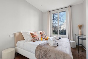 A modern hotel room with a large bed, a bedside table, a window with curtains, and a view of trees. - TheBlueStay 253 - Cozy apartment - Paris 12th (Paris)