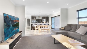 A modern living room with a flat-screen TV, a wooden coffee table, a grey sofa, and a dining area with a table and chairs. - Family Home Away From Home (Waverley)