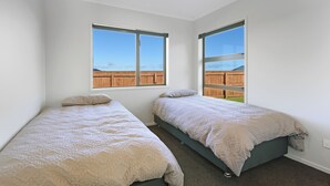 A bedroom with two beds, large windows, and a view of a wooden fence. - Family Home Away From Home (Waverley)