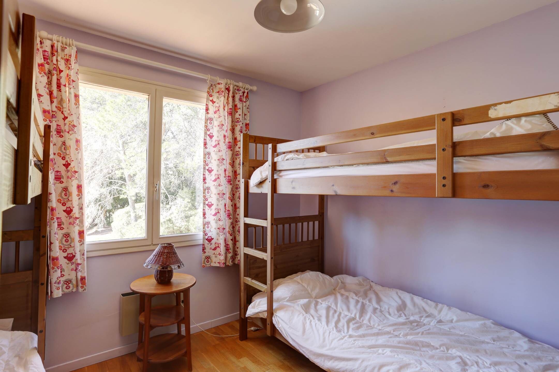 Une chambre avec un lit superposé, une fenêtre, des rideaux, une petite table avec une lampe et un radiateur.