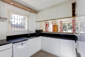 A kitchen with white cabinets, a black countertop, a dishwasher, a coffee maker, and a window with curtains. - Villa Aristide Maillol (ROQUEBRUNE SUR ARGENS)