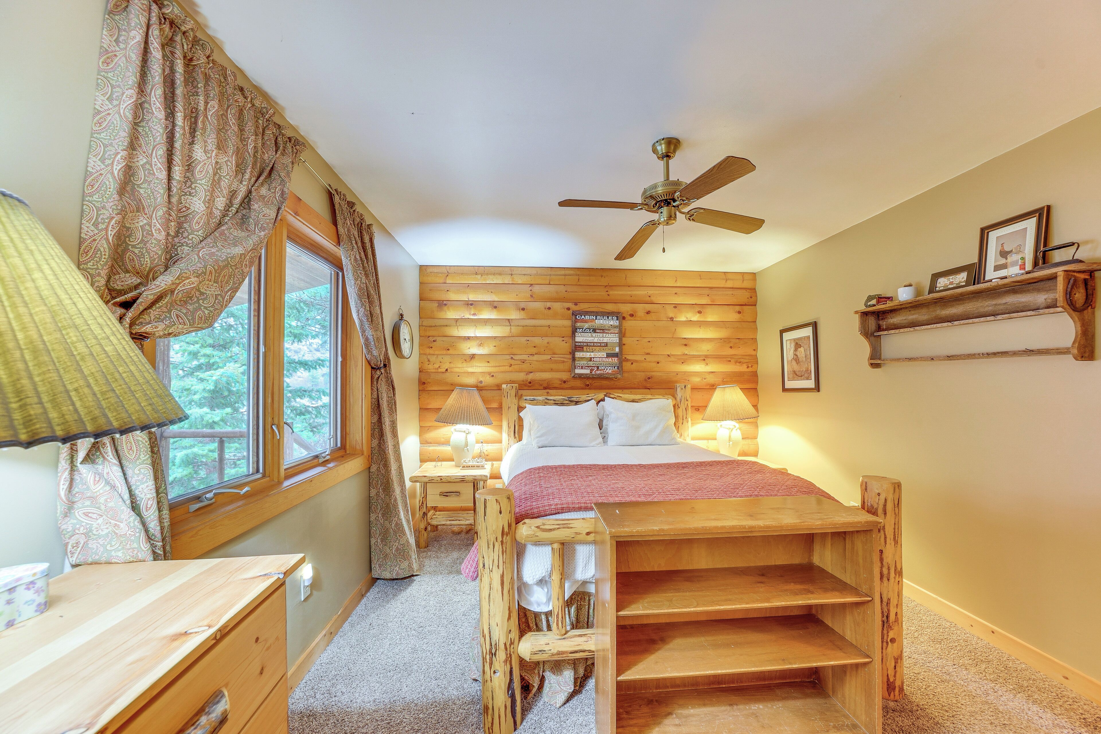 Un dormitorio acogedor con una cama de madera, una mesita de noche con lámpara, una ventana con cortinas y un ventilador de techo.