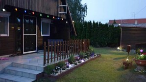 A wooden house with a front porch, steps leading to the entrance, and a garden with plants and a small fence.