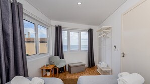 A hotel room with two beds, a desk, a chair, a small table, a bench, a wardrobe, and a view of the sea.