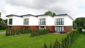 Um edifício residencial moderno de dois andares, com telhado plano, amplas janelas e um jardim bem cuidado com sebes aparadas.