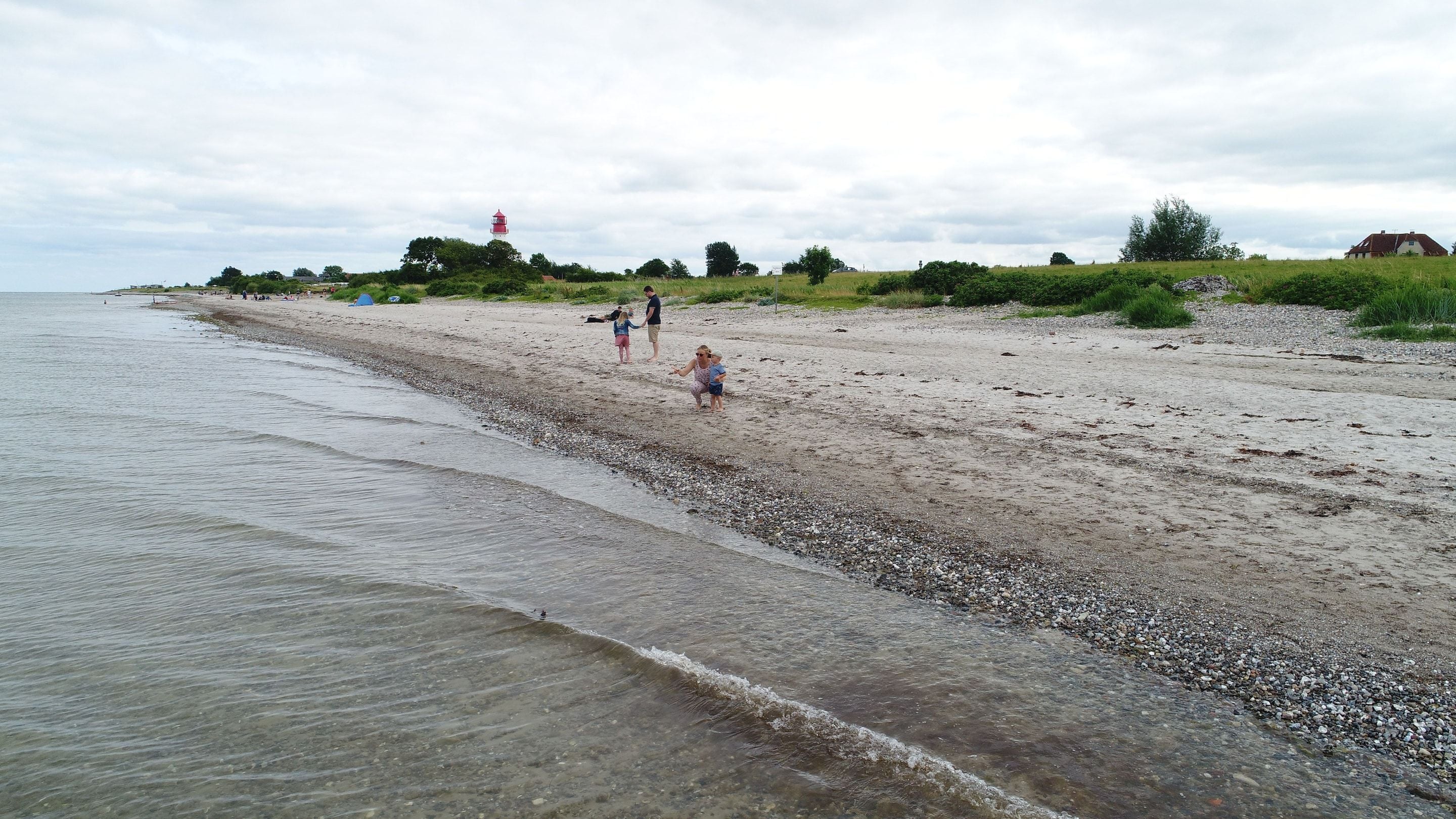 Ein kiesiger Strand mit einem Leuchtturm in der Ferne, eine begrünte Fläche mit Bäumen und ein Haus auf einem Hügel.