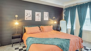 Un dormitorio con una cama, dos mesitas de noche, una mesilla auxiliar, una alfombra de cuadros y una ventana con cortinas.