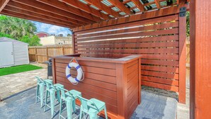 A covered outdoor patio with a wooden table and chairs, a barbecue grill, and a lifebuoy on the wall.