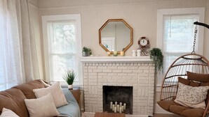 Ein Wohnzimmer mit braunem Sofa, einem Holztisch, einem Kamin aus weißen Ziegelsteinen und einem Hängesessel.