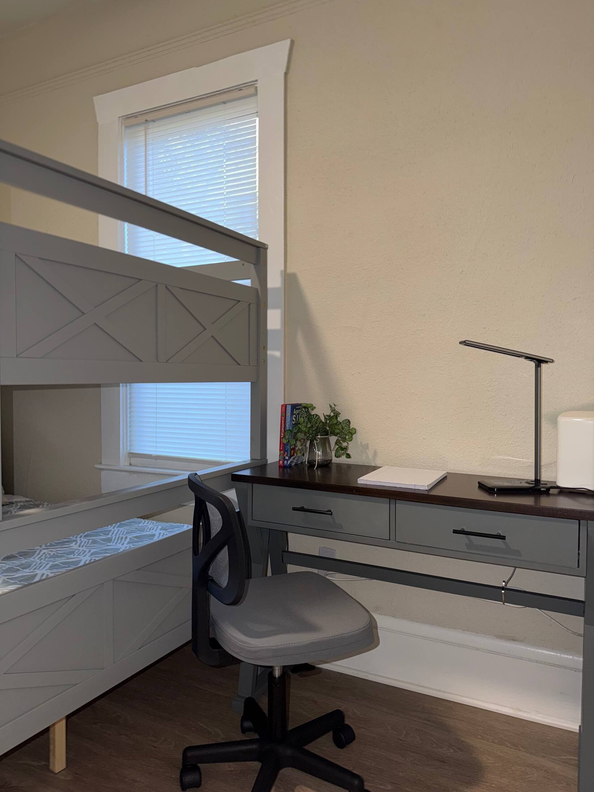 Una cama litera con escritorio y silla, una ventana con persianas, una planta y una lámpara de escritorio.