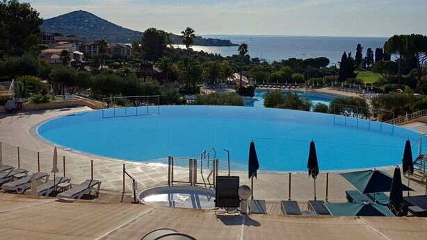 Una gran piscina azul cristalina con zonas de sol y sombrillas, rodeada de vegetación y vistas al mar y colinas distantes.