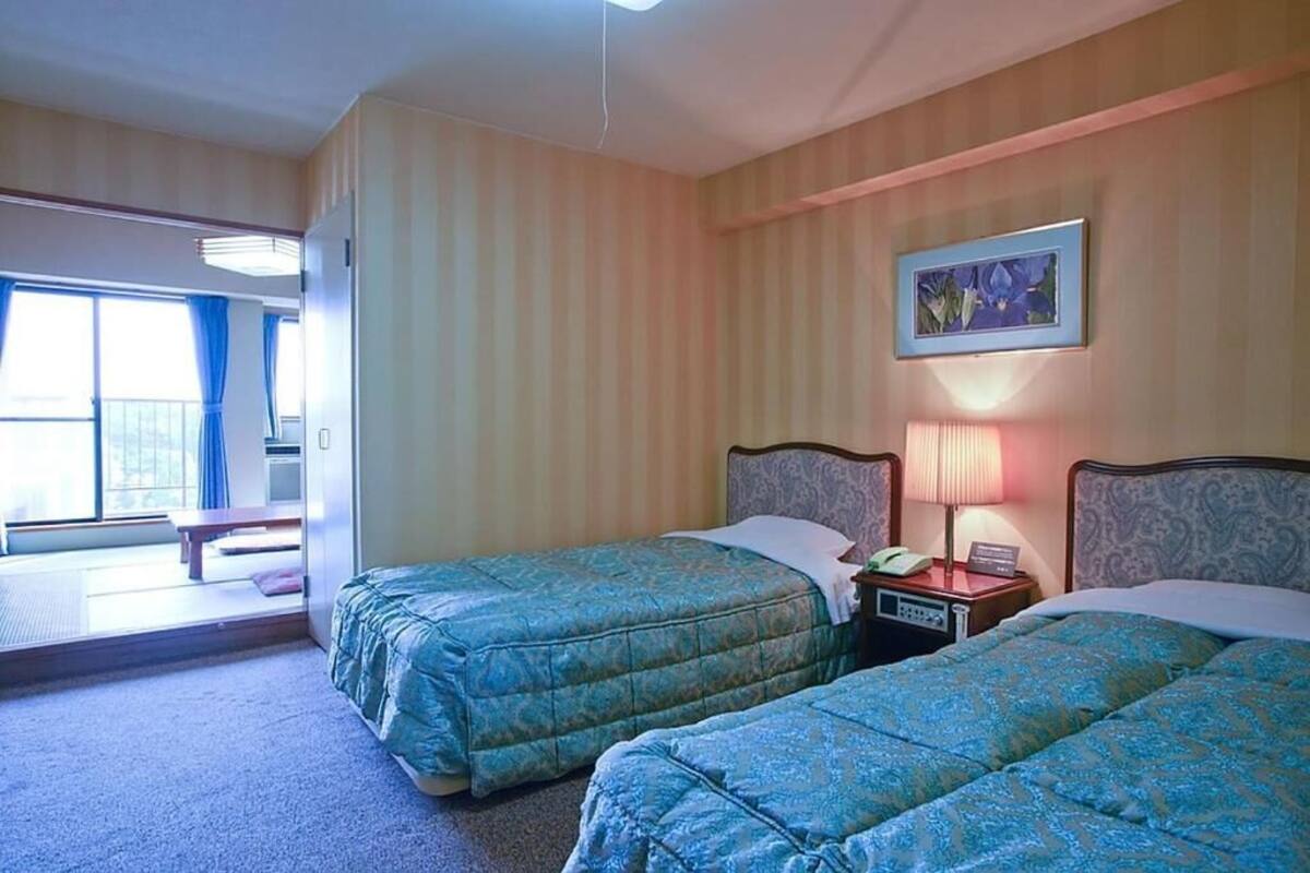 Une chambre d’hôtel avec deux lits, une table de chevet avec une lampe, un tableau au mur et une vue sur le balcon extérieur.