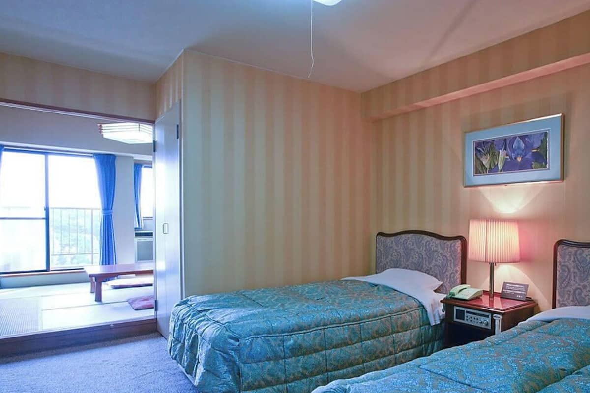 Une chambre d’hôtel avec deux lits, une table de chevet avec une lampe, un tableau au mur et une fenêtre avec des rideaux.