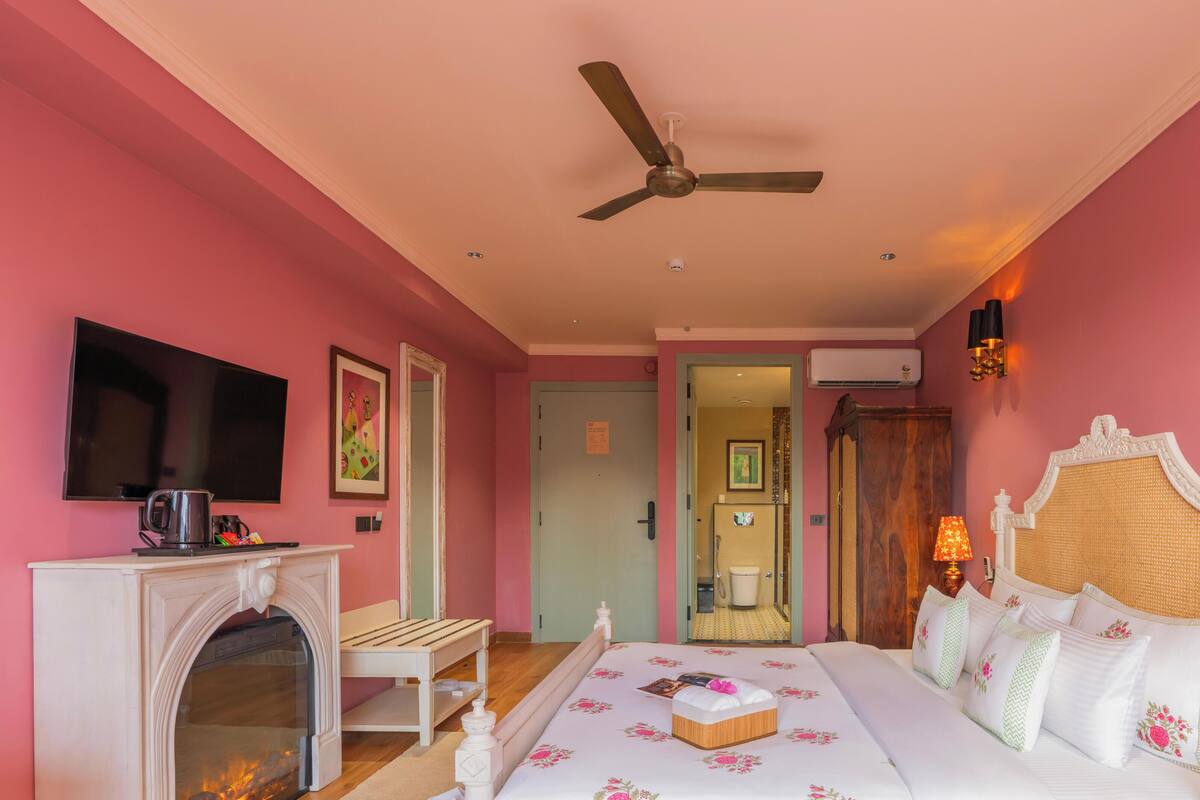 Un dormitorio con cama, televisión, chimenea, ventilador de techo y baño visible a través de una puerta abierta.