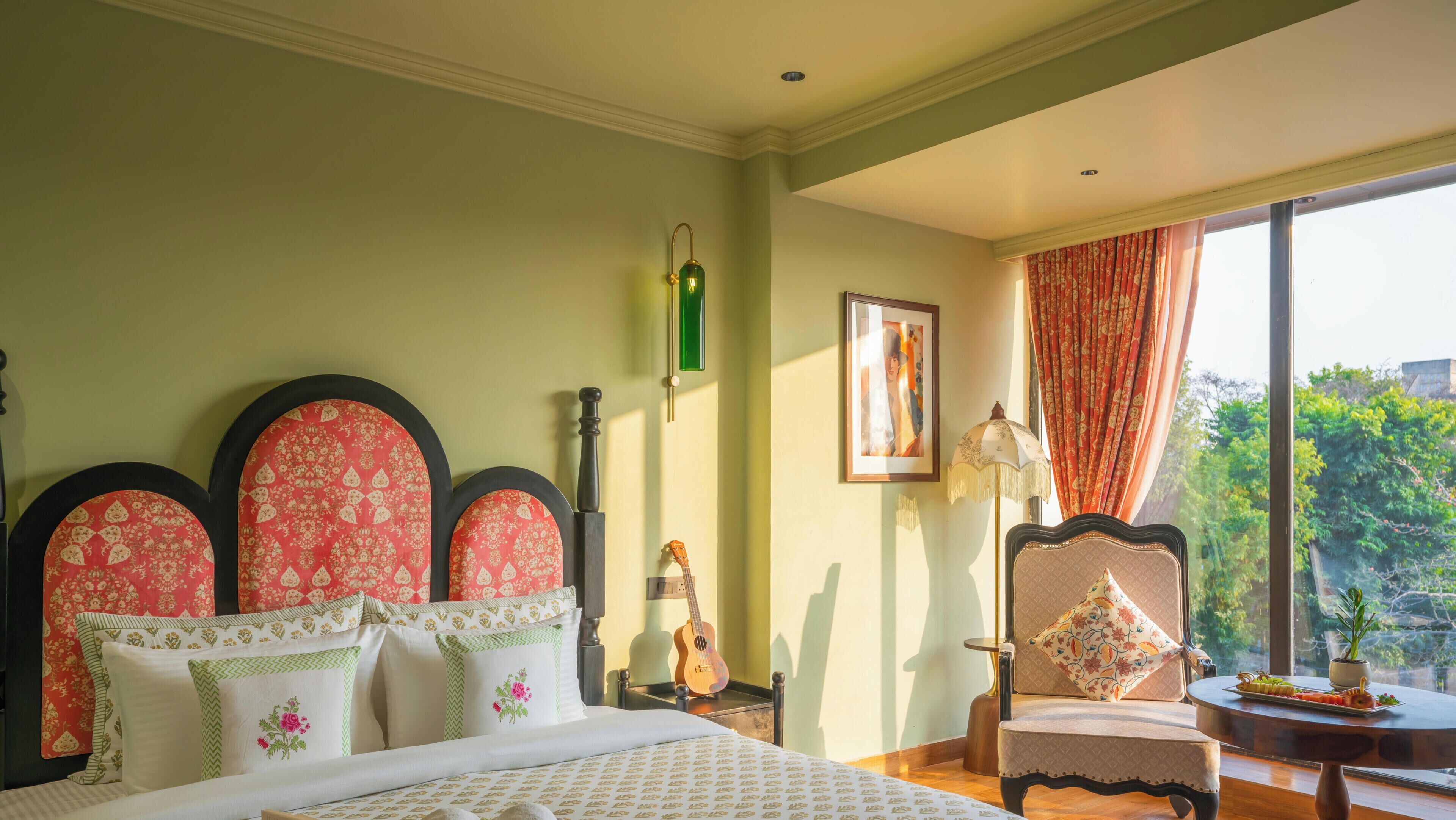 Quarto de hotel com cama, cadeira, mesa com comida, um violão e janela com vista para o verde.