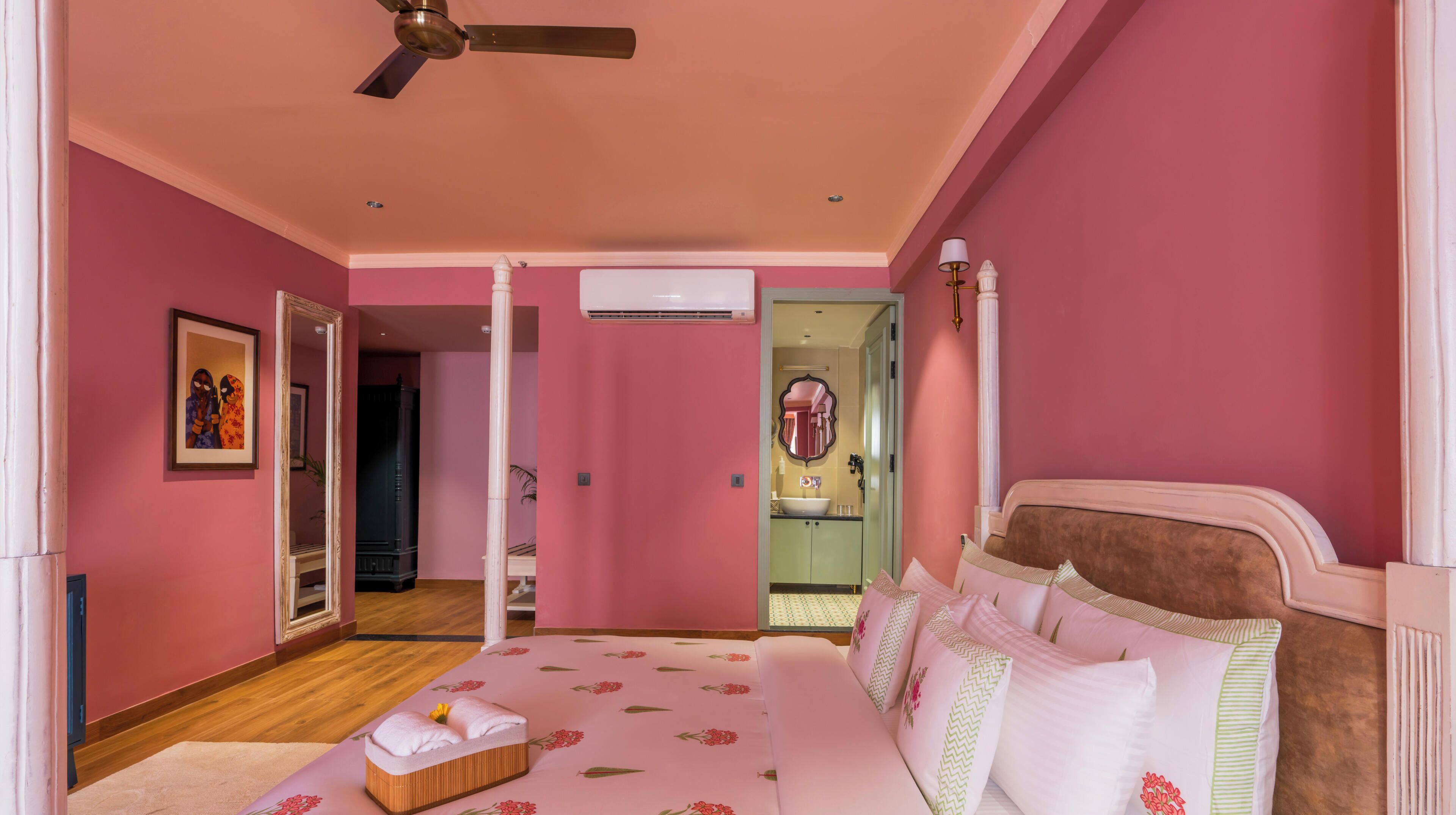 Camera da letto a tema rosa con un letto, una ventola a soffitto, uno specchio e un bagno visibile attraverso una porta aperta.