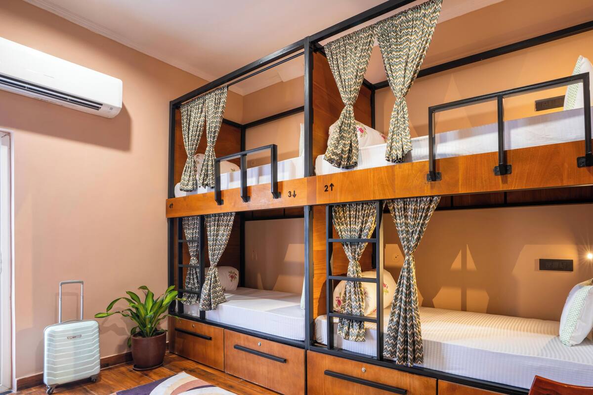 Habitación con literas de madera, cortinas, una maleta, una planta y aire acondicionado instalado en la pared.