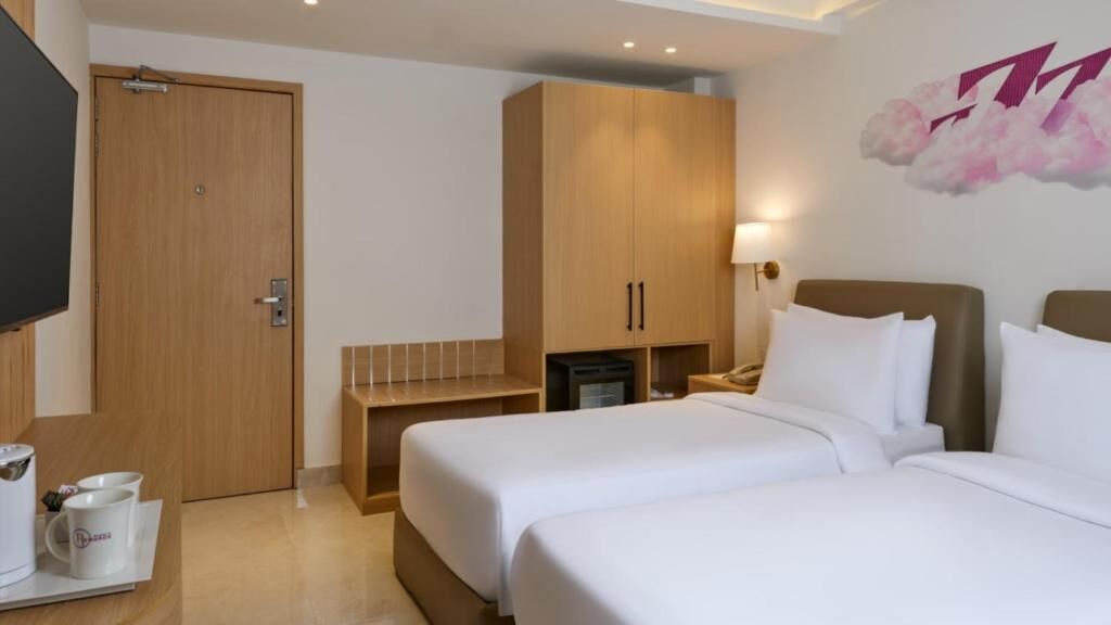 A hotel room with two beds, a wooden wardrobe, a desk with a chair, a flat-screen TV, and a wall-mounted lamp.