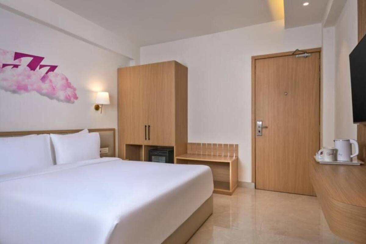 Quarto de hotel com cama, guarda-roupa de madeira, escrivaninha, televisão e luz acoplada à parede.