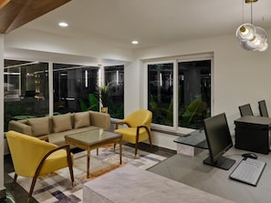 Un salon moderne comprenant un canapé, des fauteuils jaunes, une table basse, un bureau avec un ordinateur et de grandes fenêtres donnant sur un paysage verdoyant.