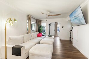 Un salon moderne avec un canapé d’angle blanc, un ventilateur de plafond, un téléviseur à écran plat et une grande fenêtre avec des rideaux.