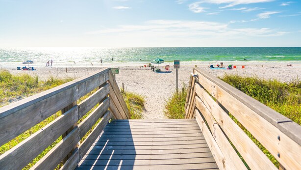 Eine Holzpromenade führt zu einem Strand mit blauem Himmel und ruhigem Wasser.
