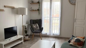 Uma sala de estar moderna com televisão de tela plana, mesa de centro, sofá, poltrona, luminária, relógio e janela com vista.