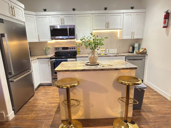 A modern kitchen with white cabinets, stainless steel appliances, a granite countertop, and two gold stools.