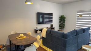 A modern living room with a dark blue sofa, a round wooden dining table with a yellow placemat, a flat-screen TV mounted on the wall, and a potted plant.