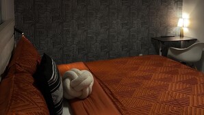 A hotel room with a patterned wallpaper, a bed with an orange bedspread, a white chair, and a desk with a lamp.