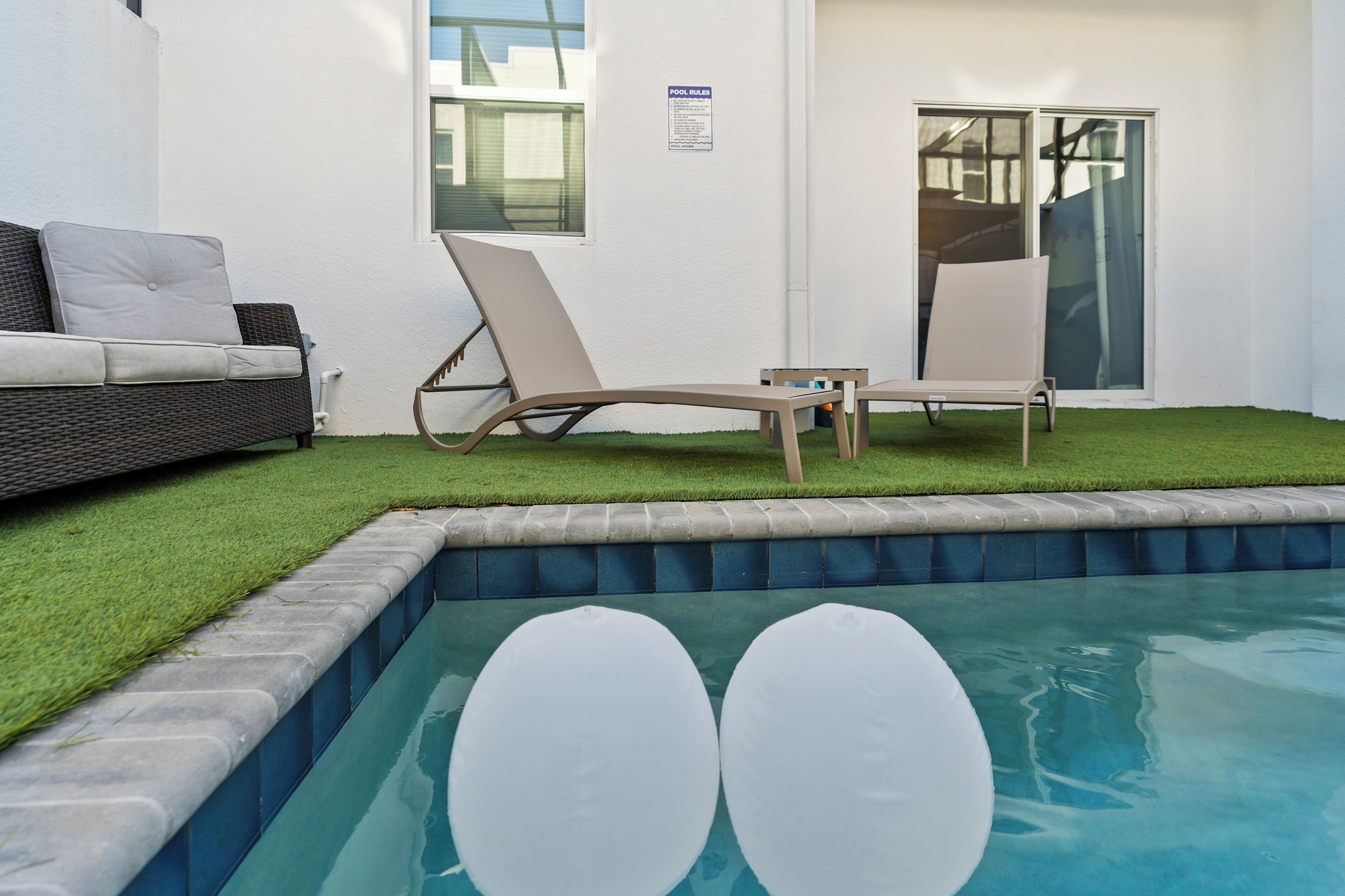 A pool area with a white pool, two lounge chairs, a white sofa, and a small patch of artificial grass.