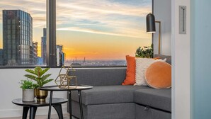 Een moderne woonkamer met een grijze bank, een kleine ronde tafel en een vloerlamp. Er is een groot raam met uitzicht op de stad.