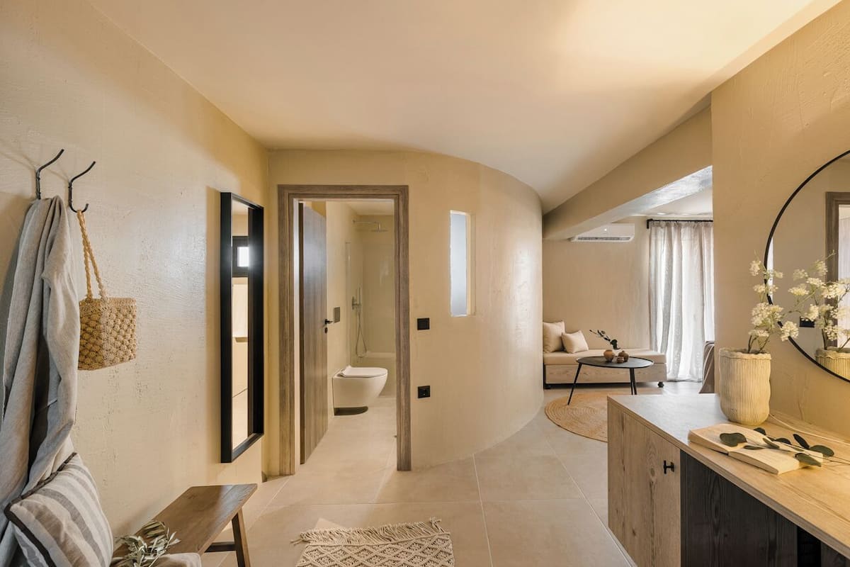 A modern interior with a curved wall, a wooden bench, a mirror, a hanging bag, and a view into a bathroom.