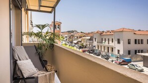 Un balcón con una silla, una planta y vista a una calle con edificios y coches aparcados.