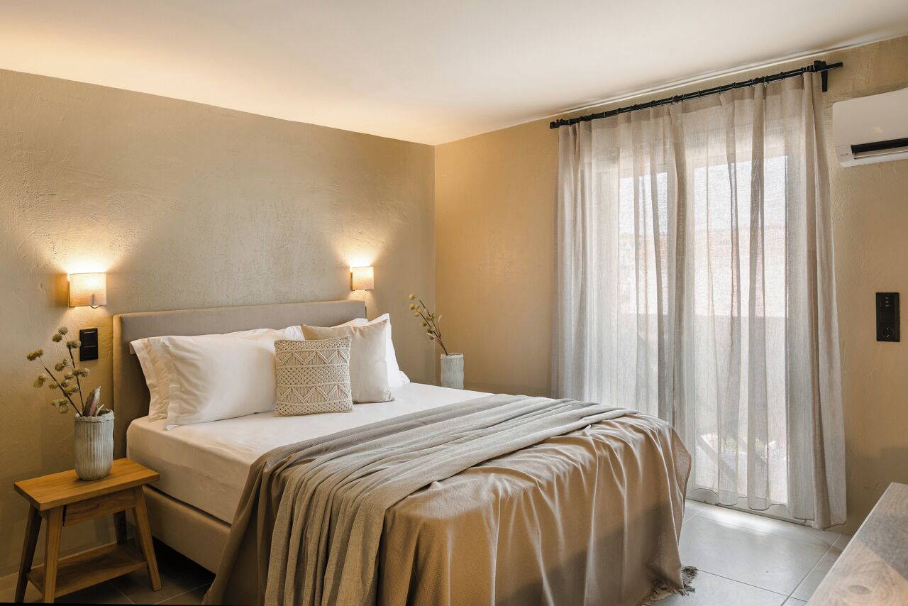 Un dormitorio con cama, lámparas de noche, mesita de noche, ventana con cortinas translúcidas y aire acondicionado de pared.