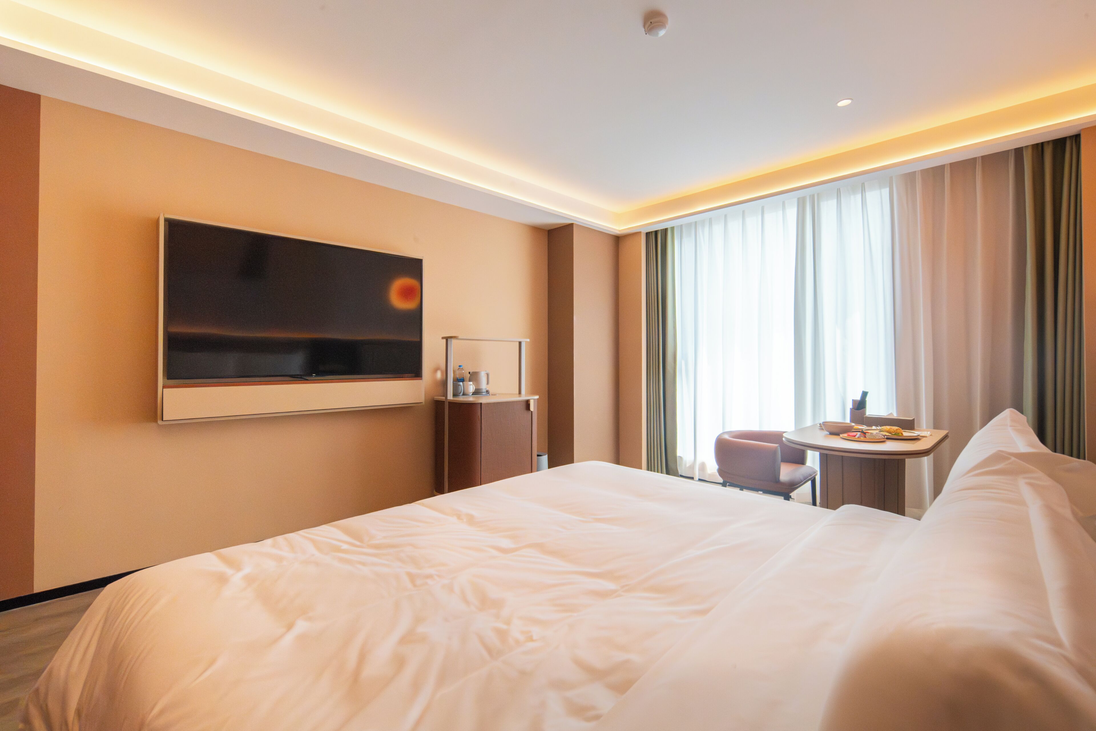 A hotel room with a large bed, a TV mounted on the wall, a small table with a chair, and a window with curtains.