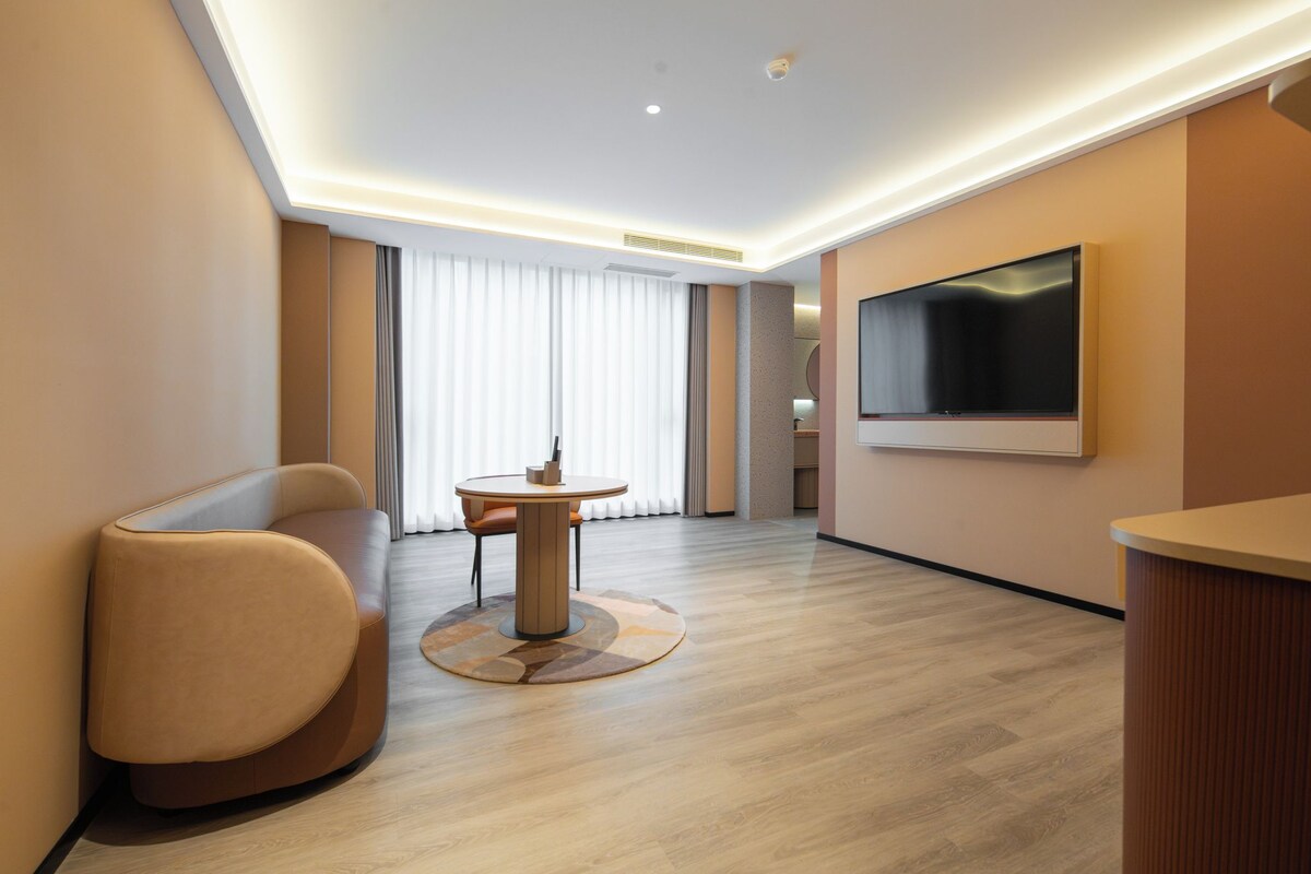 A modern hotel room with a round table, a sofa, a TV mounted on the wall, and a large window with curtains.