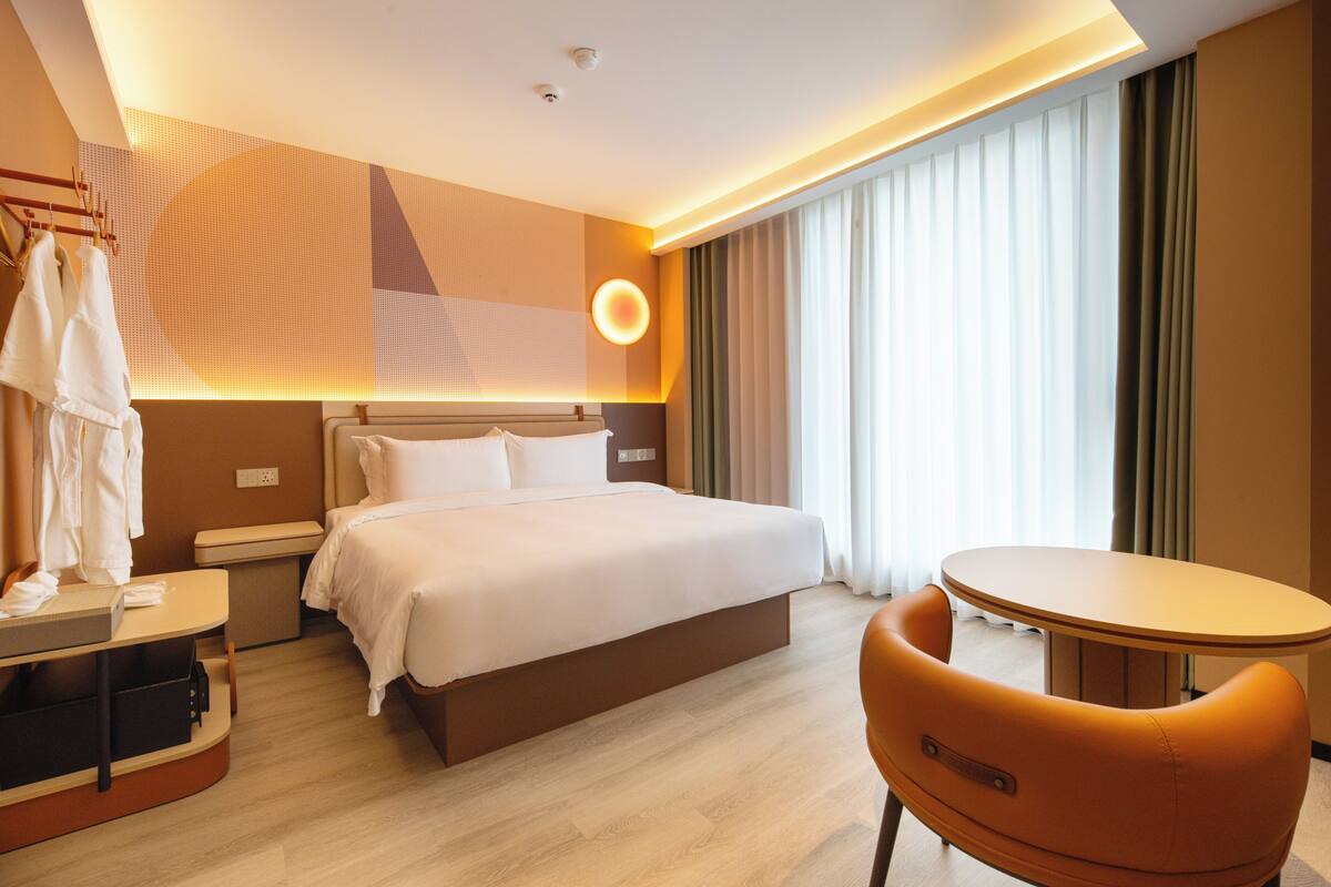 Ein Hotelzimmer mit Bett, Nachttisch, Stuhl und einem großen Fenster mit Vorhängen.