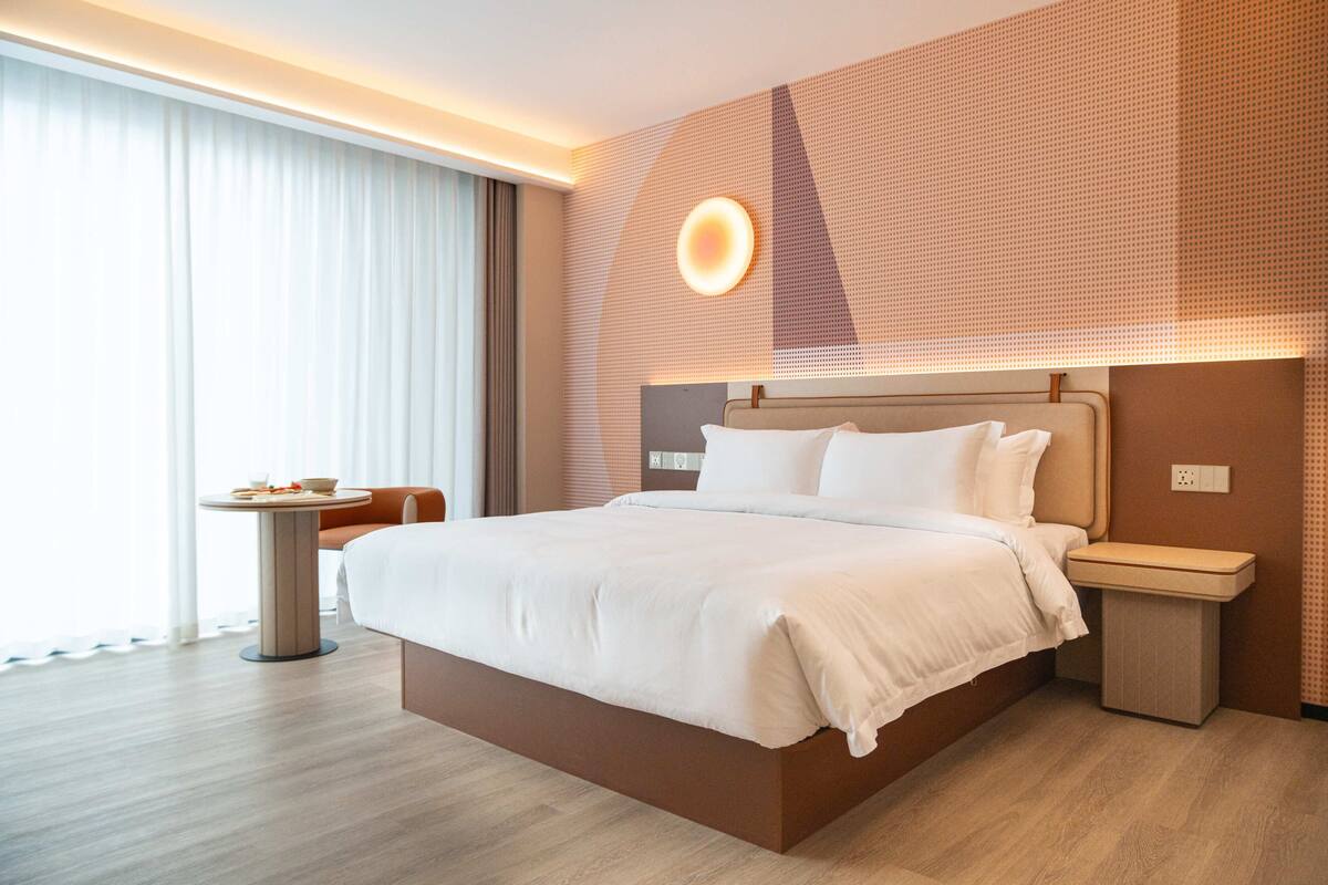 Ein modernes Hotelzimmer mit einem großen Bett, Nachttischen, einem kleinen runden Tisch und einem großen Fenster mit Vorhängen.
