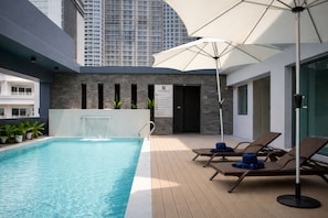 Una piscina en la azotea con sillones de descanso y sombrillas, un elemento decorativo de agua y vistas a rascacielos.
