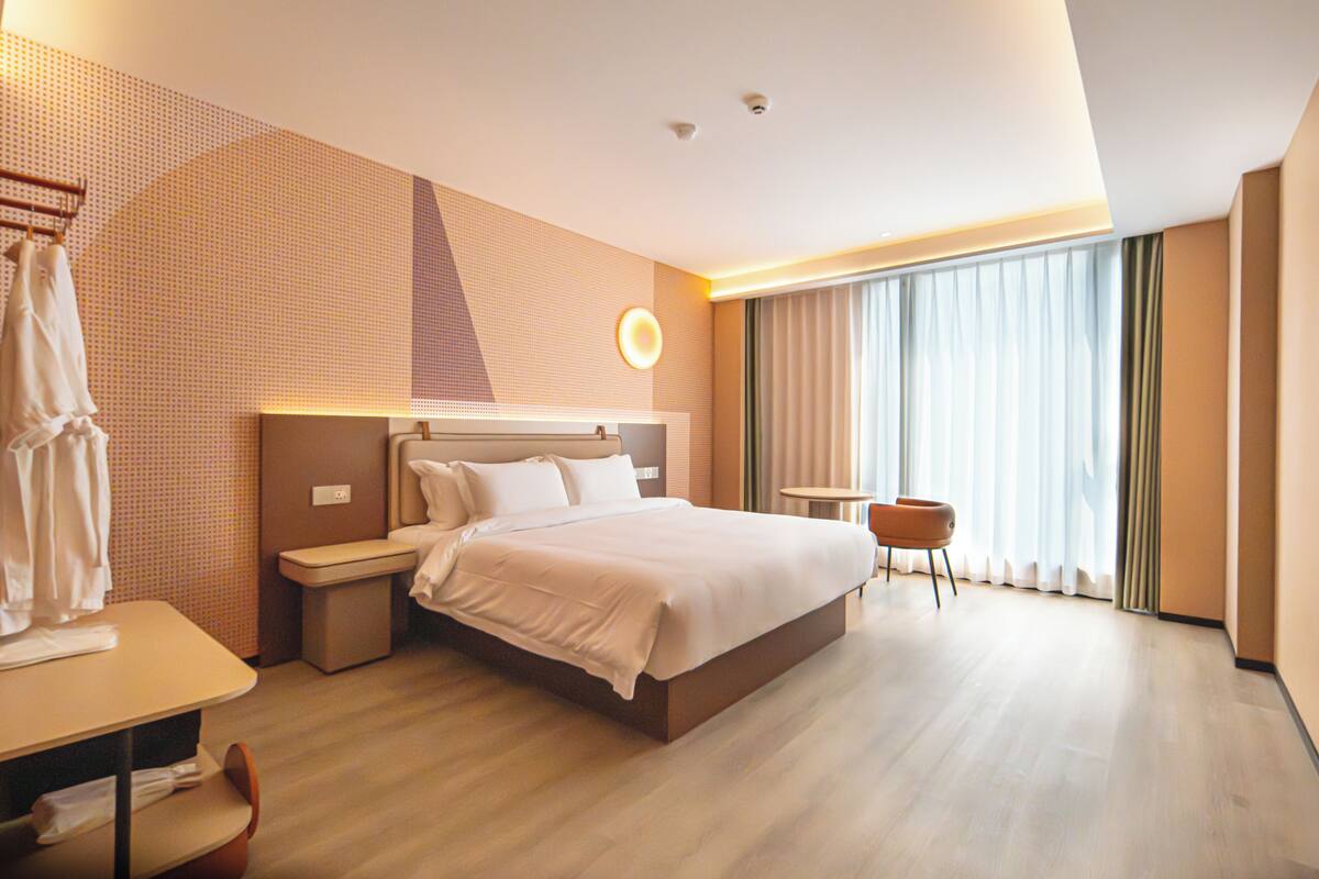 Ein modernes Hotelzimmer mit einem großen Bett, Nachttischen, einem Schreibtisch, einem Sessel und einem großen Fenster mit Vorhängen.